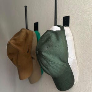 May include: Three baseball caps in shades of brown, green, and white hang on black metal wall hooks. The caps are displayed against a neutral-colored wall, showcasing a simple and organized storage solution for hats.