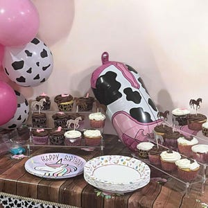 May include: A party scene with pink and cow-print balloons, cupcakes, and plates. Cupcakes are arranged on tiered stands, some topped with horse figurines. Plates feature a unicorn design and the words "Happy Birthday."