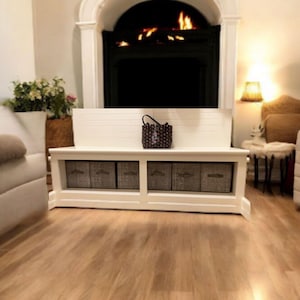 May include: A white storage bench with five gray fabric bins and a decorative handbag. The bench is in front of a fireplace with a lit fire. The room has hardwood floors and a neutral color scheme.
