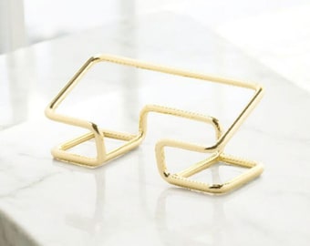 May include: A gold-colored metal wine bottle holder. The holder has a geometric design with a rectangular top and two angled supports. The holder is designed to hold a single bottle of wine. The holder is made of metal.