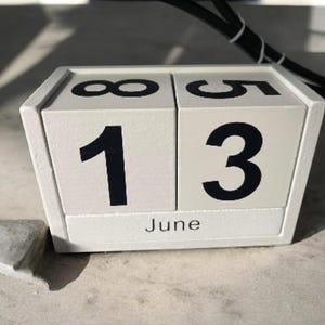 May include: A white wooden perpetual calendar displaying the date as June 13. The numbers are black and the month is printed in a simple font. The calendar is sitting on a light-colored surface.