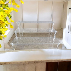 May include: Three-tiered clear acrylic display stands. Each stand has a rectangular top supported by clear legs. The stands are arranged in a stepped configuration, creating a visually appealing presentation. The background is a neutral color, allowing the stands to stand out.