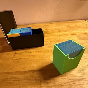 May include: A black box with a lid open, containing blue and yellow card holders filled with cards. A separate green card holder with a curved opening holds a stack of blue cards. The items are on a wooden surface.