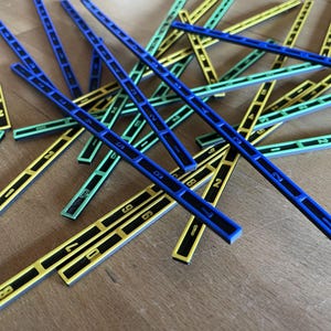 May include: A collection of colorful wooden counting sticks. The sticks are blue, yellow, and green, with black numbers printed on them. The sticks are scattered on a wooden surface.