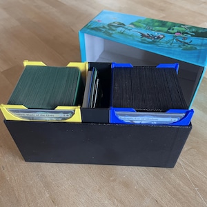 Könnte beinhalten: Eine schwarze Kartenaufbewahrungsbox mit gelben und blauen Trennwänden. Die Box enthält Kartenstapel, mit Text auf den Kartenhaltern. Der Deckel zeigt eine Unterwasserszene.