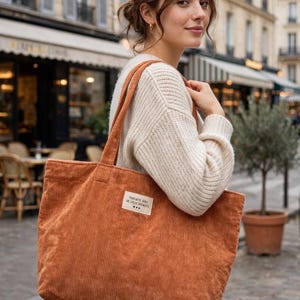 Puede incluir: Un bolso tote rectangular grande de pana color óxido con dos correas para el hombro. Una etiqueta rectangular de tela blanca está cosida en la parte delantera, con el texto negro "PARFAITE AVEC DE JOLIS DEFAUTS" y tres pequeños corazones.