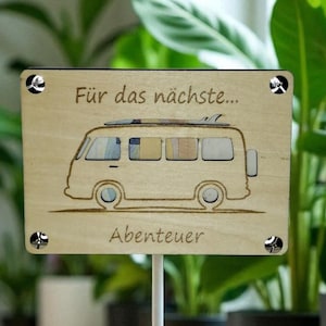Op de afbeelding: Een houten bord met de tekst "Für das nächste... Abenteuer" en een tekening van een camperbusje. Het bord is bevestigd aan een witte paal met zilveren schroeven. Op de achtergrond groene planten in zwarte potten.