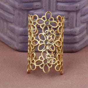 May include: A gold-colored, openwork ring featuring a floral design. The ring is made of metal and has a series of flower-shaped cutouts. The ring is a wide band, and the floral pattern repeats around the entire circumference.