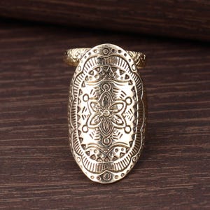 May include: A gold-coloured finger ring with an elongated oval design. The ring features intricate embossed patterns, including floral and geometric shapes. The ring is made of a shiny, metallic material and is set against a dark wood background.
