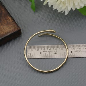 Könnte beinhalten: Ein goldfarbenes, offenes Armband mit einem schlangenartigen Design. Das Armband hat einen Durchmesser von etwa 6,35 cm. Das Armband hat ein strukturiertes Muster und wird auf einer grauen Oberfläche mit einem Lineal angezeigt.