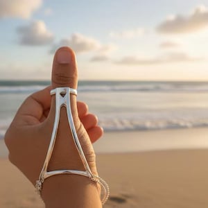 以下が含まれることがあります： シルバーの親指リングとハンドチェーン。 リングは、手の下に伸び、繊細なチェーンブレスレットに接続する三角形の形状の幾何学的なデザインが特徴です。 ジュエリーはビーチの背景に対して表示されます。