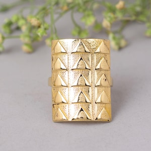 May include: A gold-colored ring with a rectangular face. The ring's face is textured with a repeating pattern of small, raised triangles. The ring is set against a neutral background with a blurred floral element.