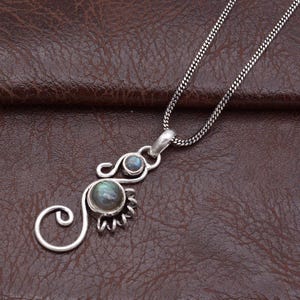 May include: A silver pendant necklace with a swirling design, two small blue gemstones, and a larger, round labradorite stone. The necklace has a delicate chain. The background is brown leather.