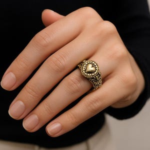 May include: A gold-colored ring featuring a heart-shaped design at the center, surrounded by a circular pattern. The ring has intricate detailing and is worn on a finger. The ring's band has a decorative pattern.