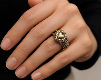 Puede incluir: Un anillo dorado con un diseño en forma de corazón en el centro, rodeado por un patrón circular. El anillo tiene detalles intrincados y se lleva en un dedo. La banda del anillo tiene un patrón decorativo.