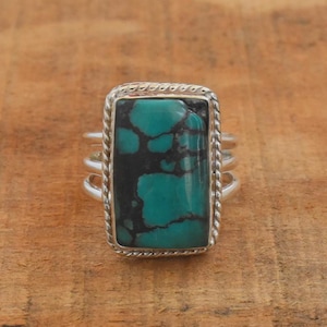 Puede incluir: Un anillo de plata con una piedra rectangular de turquesa con patrones de matriz negra. La piedra está engastada en un bisel estilo cuerda. El anillo tiene un diseño de triple banda.