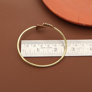 May include: A gold-coloured bangle bracelet with a geometric design. The bracelet is circular, with a triangular accent and a series of small, linear details. The bracelet is approximately 6.35 cm in diameter. The words "MADE IN INDIA" are visible.