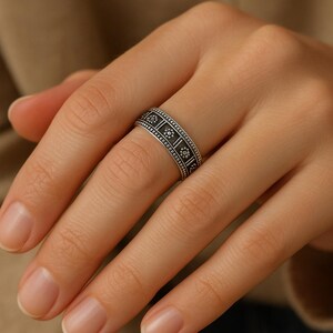 May include: A silver ring with a repeating pattern of square and floral designs. The ring is worn on a finger, showcasing the intricate details of the jewelry. The ring's design is detailed with small dots.