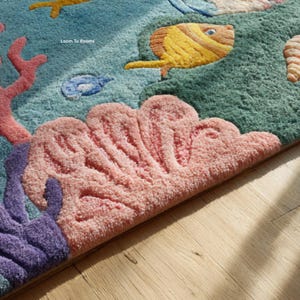 Puede incluir: Primer plano de una alfombra colorida y texturizada con un tema submarino. La alfombra presenta corales, peces y otras criaturas marinas en tonos rosa, amarillo, azul y verde. La alfombra est&aacute; colocada sobre un suelo de madera.