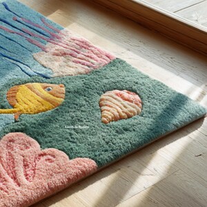 Puede incluir: Una alfombra rectangular con un dise&ntilde;o de escena submarina. La alfombra presenta un pez amarillo, una concha y coral en tonos de rosa, verde y azul. El texto "Loom To Rooms" es visible en la alfombra.