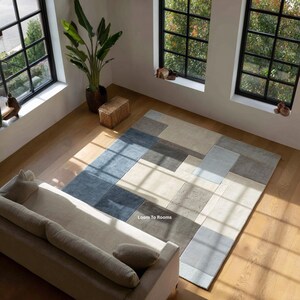 Puede incluir: Una alfombra rectangular con un patrón geométrico en tonos azules, grises y beige. La alfombra está colocada sobre un suelo de madera, con un sofá y ventanas en el fondo. El texto "Loom To Rooms" es visible en la alfombra.