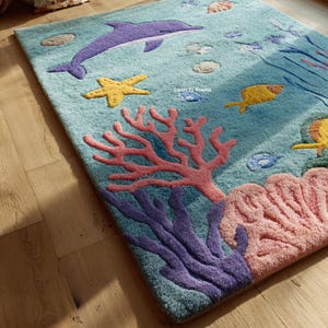 Puede incluir: Una alfombra rectangular con tem&aacute;tica submarina. La alfombra presenta un delf&iacute;n morado, una estrella de mar amarilla y coral en tonos rosa y morado sobre un fondo azul claro. Tambi&eacute;n se representan peque&ntilde;os peces y otras criaturas marinas.