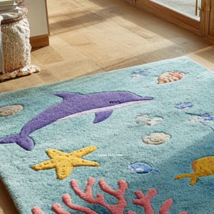 Puede incluir: Una alfombra rectangular azul claro con tem&aacute;tica marina. Un delf&iacute;n morado est&aacute; centrado, rodeado de estrellas de mar amarillas, corales y varias criaturas marinas. La alfombra tiene un aspecto suave y texturizado, adecuada para la habitaci&oacute;n de un ni&ntilde;o o el ba&ntilde;o.