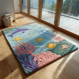 Puede incluir: Alfombra rectangular con una escena submarina. Presenta un delf&iacute;n morado, una estrella de mar amarilla y corales coloridos. El fondo es azul claro, con peces y conchas. La alfombra est&aacute; hecha de un material suave y afelpado.
