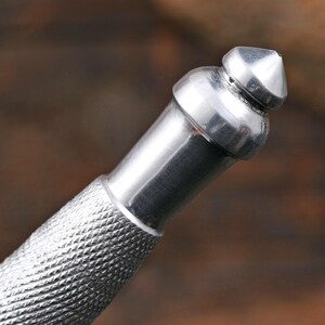 May include: A silver-colored metal tool handle featuring a textured, cross-hatched knurled grip section that transitions into a smooth, cylindrical neck and terminates in a pointed, dome-shaped finial at the very top of the object.