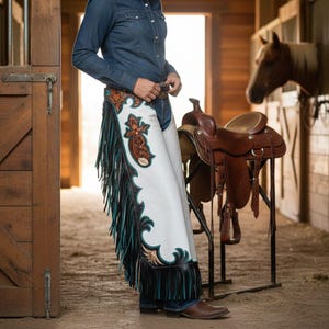 May include: White leather chaps with turquoise and black fringe and decorative brown and turquoise accents. The chaps have a floral design and are worn with a denim shirt and jeans. A brown leather saddle is in the background.