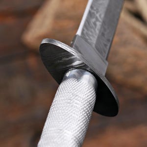 May include: A knife with a silver-colored, textured, diamond-patterned metal handle and a dark, patterned Damascus steel blade. The knife features a dark, oval-shaped metal guard separating the handle from the blade, showing visible metal layers.