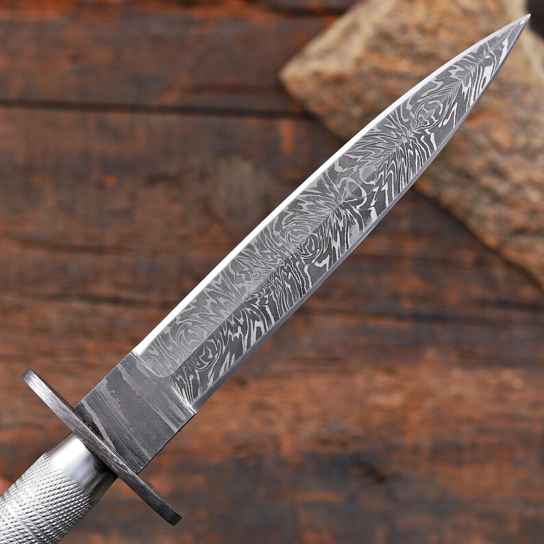 May include: A double-edged dagger featuring a pointed blade with a complex, swirling gray and silver Damascus steel pattern. The blade connects to a silver-toned metal crossguard and a textured, cylindrical handle with a cross-hatched grip pattern.