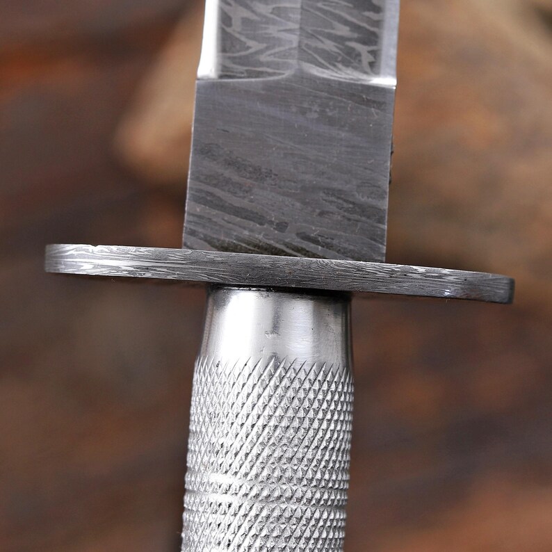 May include: A knife featuring a Damascus steel blade with a distinct wavy pattern, a flat metal crossguard, and a cylindrical handle with a textured, diamond-patterned knurled grip. The blade and guard exhibit a dark, layered metallic finish.