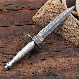 May include: A double-edged dagger featuring a pointed blade with a distinct wavy Damascus steel pattern. The handle is made of silver-toned metal with a textured, cross-hatched knurled grip and a rounded pommel, separated by a small metal crossguard.