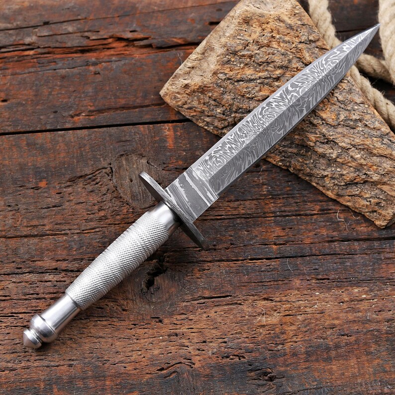 May include: A double-edged dagger featuring a pointed blade with a distinct wavy Damascus steel pattern. The handle is made of silver-toned metal with a textured, cross-hatched knurled grip and a rounded pommel, separated by a small metal crossguard.