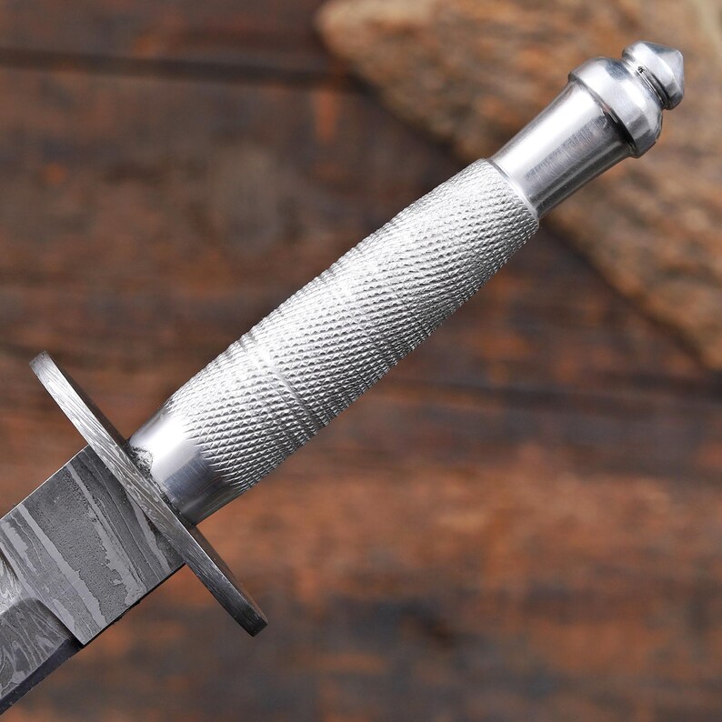 May include: A silver-colored metal knife handle featuring a textured, diamond-patterned grip and a smooth, rounded pommel. The handle connects to a flat, dark metal crossguard and a blade displaying a distinct, wavy, layered pattern throughout.