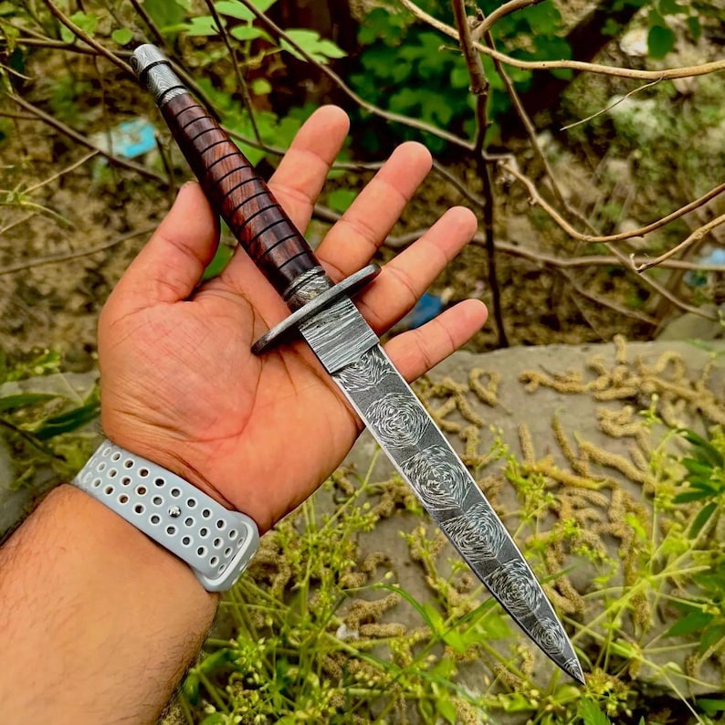 May include: A double-edged dagger featuring a dark brown, ribbed wooden handle and a silver-toned metal crossguard. The blade is crafted from patterned Damascus steel, displaying a distinct swirling, layered metal design that tapers to a point.