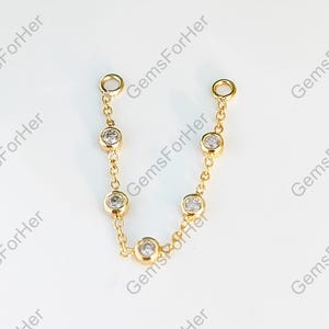 May include: A gold chain featuring seven clear, round gemstones. The chain is composed of bezel-set stones linked together. The jewellery is presented against a white backdrop. The text "GemsForHer" is visible. The chain is a delicate piece.