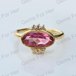 Puede incluir: Un anillo de oro con una gema rosa ovalada y facetada. El anillo está adornado con pequeñas gemas redondas y transparentes en la parte superior e inferior del óvalo. El anillo está sobre un fondo blanco, con el texto "Gems For Her" en el fondo.