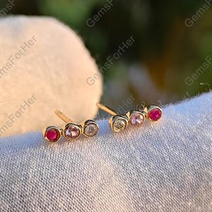 Pode incluir: Um par de brincos de ouro, cada um com três pedras preciosas: uma rubi, uma gema rosa e uma gema transparente. Os brincos são exibidos em uma superfície branca, com o logotipo GemsForHer visível ao fundo.