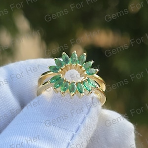 Könnte beinhalten: Ein goldener Ring mit floralem Design und smaragdgrünen Edelsteinen. Der Ring hat ein Doppelband und einen offenen Mittelkreis. Der Ring wird vor einem weißen Hintergrund mit dem Text "Gems For Her" gehalten.