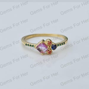 May include: A gold ring with a central square, pink gemstone, complemented by orange and blue stones. The band is decorated with small green gemstones. The ring is set against a white background.
