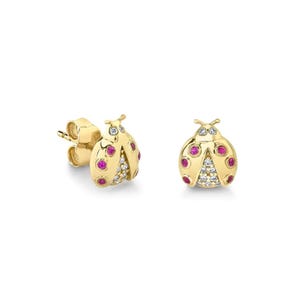 May include: A pair of gold-colored ladybug stud earrings. Each ladybug has red and white gemstone accents. The earrings are set against a white background.