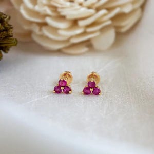 Puede incluir: Un par de pendientes de oro con tres gemas magenta redondas dispuestas en un racimo triangular. Los pendientes están sobre un fondo blanco suave, con un elemento floral borroso en el fondo. Las gemas son de color intenso.