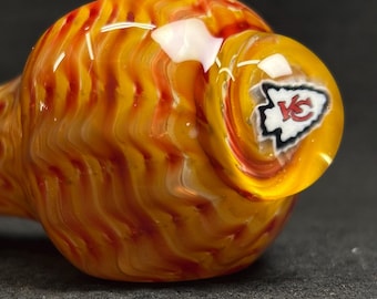 May include: A hand-blown glass pipe with a swirling pattern of orange, yellow, and red. The bowl features the Kansas City Chiefs logo in white and red on a yellow background. The pipe is designed for smoking.