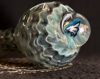 Puede incluir: Una pipa de vidrio soplado a mano con un patrón arremolinado de colores turquesa, negro y transparente. El cuenco presenta un globo de vidrio transparente con un logotipo de águila azul y blanco. La pipa está diseñada para fumar.