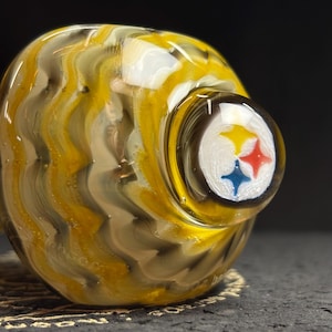 May include: A glass art piece with a swirling pattern of yellow, white, and brown. The end features the Pittsburgh Steelers logo in white, yellow, red, and blue. The piece is on a dark surface.