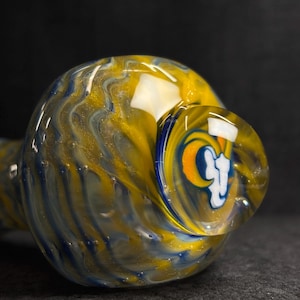 May include: A glass pipe with a swirling pattern of yellow, blue, and white. The bowl features a logo with a white ram's head on a blue and orange background. The pipe is designed for smoking.
