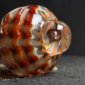 May include: A hand-blown glass pipe with a swirling pattern of red, brown, and white. A small, clear bubble on the side features a pirate skull and crossbones design. The glass has a glossy finish and is set against a dark background.