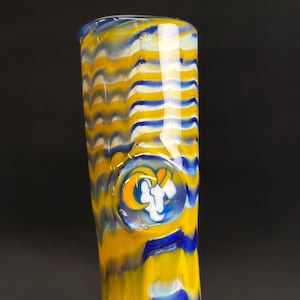 May include: A glass smoking pipe with a yellow, blue, and white wavy pattern. The pipe features a team logo in a circular design. The colors are vibrant, and the glass appears to be hand-blown.